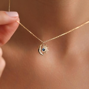 Puede incluir: Collar dorado con un colgante de luna y sol. El sol tiene un centro de piedra preciosa azul. El collar está sobre una cadena delicada. El colgante mide aproximadamente 2 cm de diámetro.