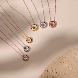 May include: A collection of gold, silver, and rose gold necklaces with gemstone pendants. The pendants feature various colored gemstones, including green, pink, blue, and clear stones. The necklaces are displayed on a light-colored surface.