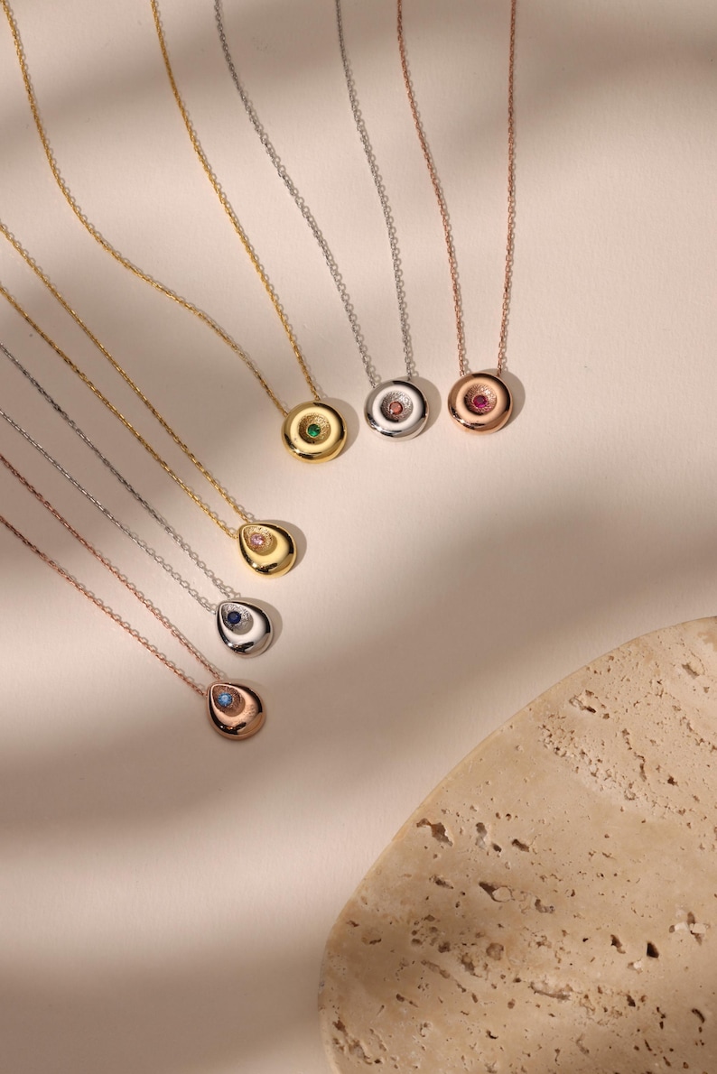 May include: A collection of gold, silver, and rose gold necklaces with gemstone pendants. The pendants feature various colored gemstones, including green, pink, blue, and clear stones. The necklaces are displayed on a light-colored surface.