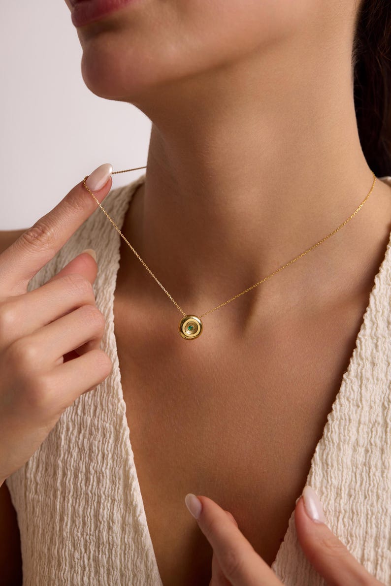 May include: A delicate gold necklace featuring a small, round pendant with a central green gemstone. The pendant is encircled by a gold bezel, and the chain is fine and elegant. The necklace is displayed on a person's neck.