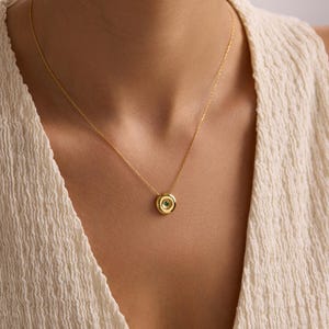 May include: A gold-colored necklace with a circular pendant. The pendant has a small, round, green gemstone at its center, surrounded by a gold-colored frame. The necklace is displayed against a neutral background.