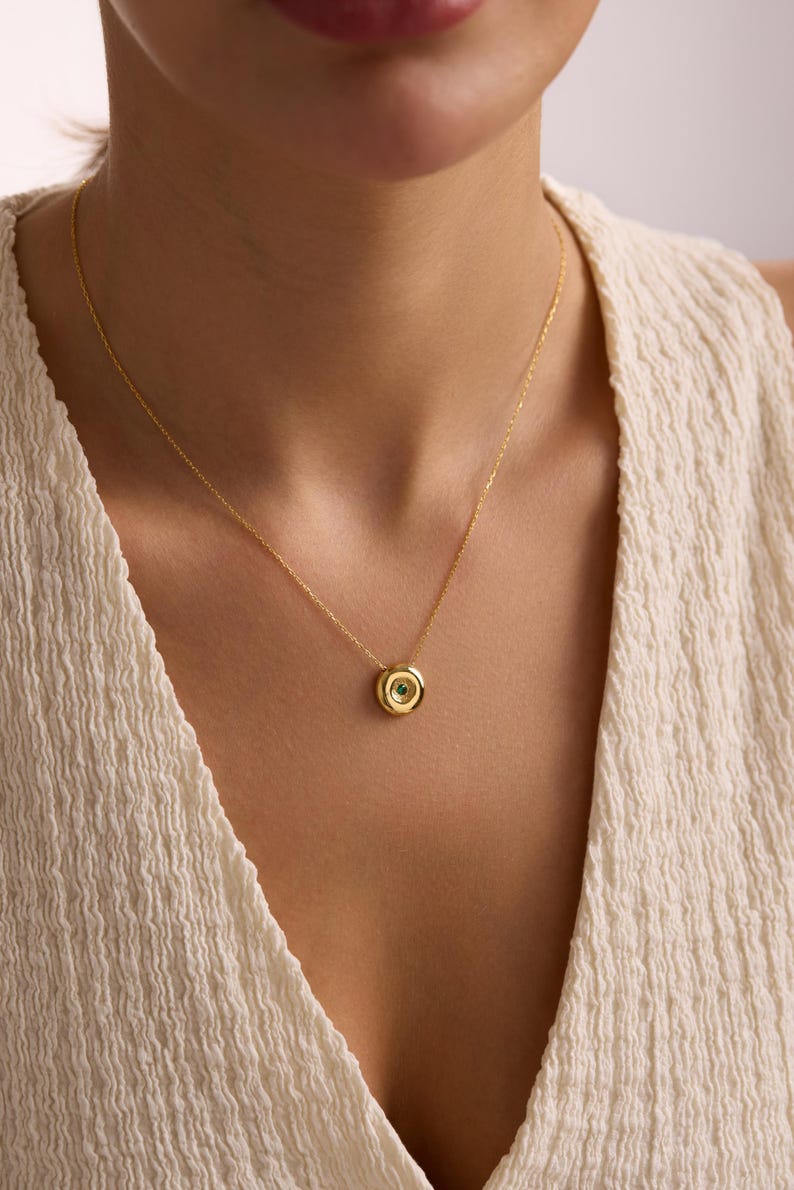 May include: A gold-colored necklace with a circular pendant. The pendant has a small, round, green gemstone at its center, surrounded by a gold-colored frame. The necklace is displayed against a neutral background.