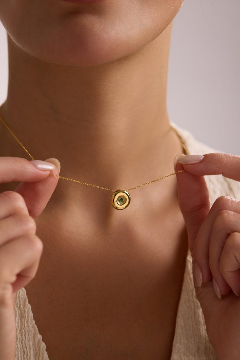 May include: A gold-colored necklace with a small, circular pendant. The pendant features a green gemstone set in the center. The necklace is being held by a person, showcasing the delicate chain and the pendant's design.