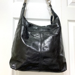 May include: A black hobo bag with a crinkled, glossy texture. The bag features a single shoulder strap attached with silver-toned metal hardware, including lobster claw clasps. A vertical seam runs down the center of the front panel.
