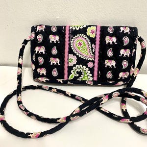 May include: Black crossbody bag with a repeating pattern of pink elephants and paisley designs. The bag features a pink and green paisley stripe down the center and a matching strap. The bag is approximately 8 inches wide.