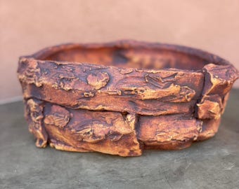 Op de afbeelding: Een rustieke, handgemaakte keramische plantenbak met een getextureerd, gelaagd ontwerp in tinten bruin en oranje. De plantenbak heeft een brede, ronde opening en een licht ongelijke rand.