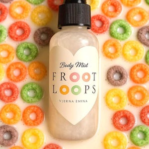 Op de afbeelding: Een heldere body mist fles met een zwarte sproeikop, met het label "Body Mist FROOT LOOPS" in een hartvormig ontwerp. De fles is omgeven door kleurrijke ontbijtgranen in een melkachtige vloeistof, wat een levendige, speelse esthetiek creëert.