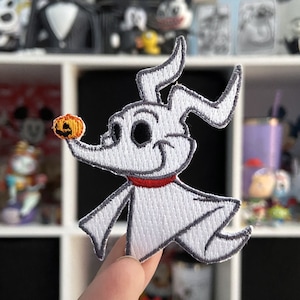 May include: Embroidered patch featuring Zero from The Nightmare Before Christmas. The white ghost dog with a red collar is holding a small orange pumpkin. The patch has a gray outline and is on a white background.