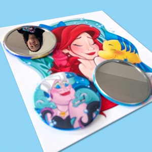 May include: Set of three round compact mirrors featuring characters from the Disney movie The Little Mermaid. The mirrors are decorated with Ariel, Ursula, and Flounder. The mirrors are surrounded by a blue and green ocean background.