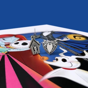 May include: A pair of silver-tone earrings featuring a black and white heart with a bat design and a black and white house with the word "ZERO" on it. The earrings are on a background of a cartoon image of Jack Skellington and Sally from the Nightmare Before Christmas.
