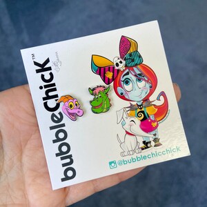 May include: A set of four enamel pins featuring characters from the Nightmare Before Christmas. The pins are colorful and feature a pink and purple dragon, a green dragon, a white dog, and a blue-skinned girl with a pink and black bow. The pins are on a white background with the text "bubblechick" and a social media handle.