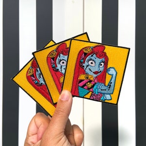 Puede incluir: Parche bordado con un personaje de dibujos animados con piel azul, cabello rojo y apariencia cosida. El personaje flexiona un bíceps. El parche tiene un fondo amarillo y un borde negro. Los parches se sostienen en una mano.