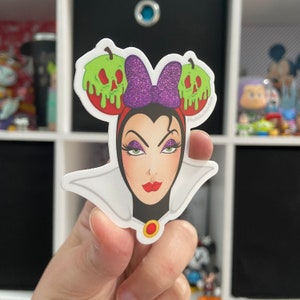 May include: A white sticker with a cartoon image of the Evil Queen from Snow White. She is wearing a purple and black headband with green apples with skulls on them. The sticker has a white collar and a gold necklace.