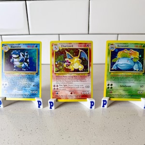 May include: Three collectible trading cards featuring Blastoise, Charizard, and Venusaur, each displayed on a small white plastic stand with a blue letter P on the sides. The cards show detailed character illustrations, HP values, and game text.
