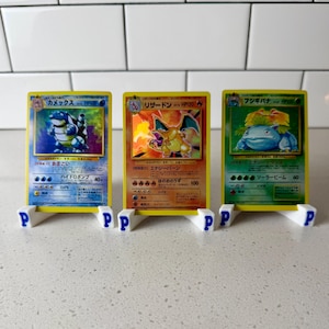 Puede incluir: Tres cartas coleccionables de Pokémon exhibidas en soportes blancos y azules. Las cartas presentan ilustraciones coloridas de personajes Pokémon. Los soportes tienen una base blanca con una "P" azul en cada uno. Las cartas están en marcos amarillos.