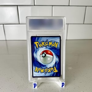 Może przedstawiać: Oceniona karta kolekcjonerska Pok&eacute;mon w przezroczystym plastikowym etui. Karta przedstawia logo Pok&eacute;mon i wz&oacute;r Pok&eacute; Ball na niebieskim tle. Etui jest podparte dwoma małymi białymi stojakami z niebieskimi literami "P".