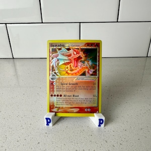 May include: A Gyarados Pokémon trading card, displayed on a white stand. The card features a red and yellow illustration of Gyarados, with text describing its abilities. The stand is white with blue "P" letters.