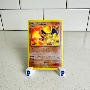 May include: A Charizard Pokémon trading card, featuring a fiery illustration of the creature. The card is yellow with red and orange accents, and is displayed on a white stand with blue "P" letters.