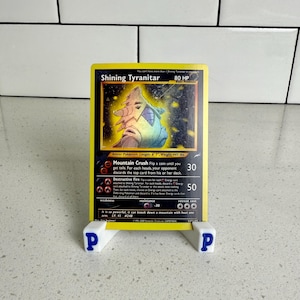Könnte beinhalten: Eine gelbe Shining Tyranitar Pokémon-Karte, präsentiert auf einem weißen Ständer mit blauen "P"-Buchstaben. Die Karte zeigt eine farbenfrohe Illustration eines Wesens und Text, der seine Fähigkeiten beschreibt. Die Karte ist 201 cm lang und wiegt 201 kg.
