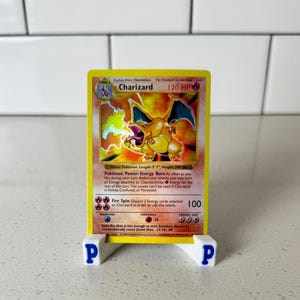 May include: A Charizard Pokémon card, displayed on a white stand with blue lettering. The card features a yellow border and an illustration of a fiery dragon. Text on the card includes "Charizard" and "120 HP".