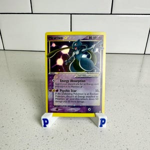 Puede incluir: Una carta coleccionable de Pokémon con Mewtwo, una criatura morada y blanca, con el texto "Mewtwo" y "80 HP". La carta tiene un borde amarillo y se muestra en un soporte blanco con la letra "P" a cada lado.