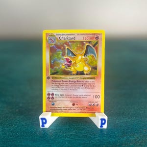 Peut inclure: Une carte à collectionner Charizard avec une bordure jaune, une illustration de type feu et 120 PV, présentée sur un support en plastique blanc avec des lettres P bleues sur les côtés. Le texte inclut Pokémon Power Energy Burn et Fire Spin.