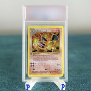 Puede incluir: Una carta de Pokémon Charizard en una funda protectora, exhibida en un soporte blanco. La carta presenta un fondo amarillo con una ilustración de Charizard y texto. La carta está clasificada y encerrada en un soporte de plástico transparente.
