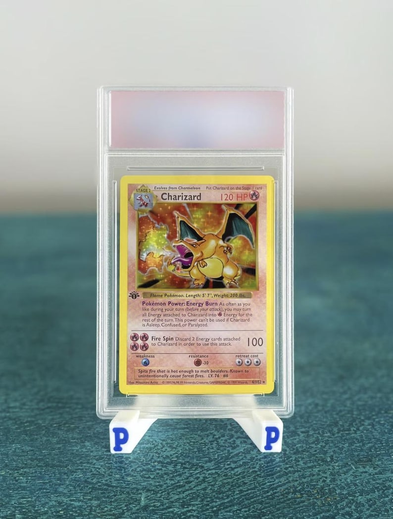 May include: A Charizard Pok&eacute;mon card in a protective case, displayed on a white stand. The card features a yellow background with a Charizard illustration and text. The card is graded and encased in a clear plastic holder.