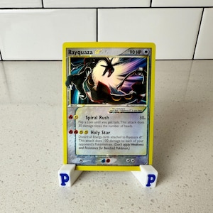 May include: A Rayquaza Pokémon card with a yellow border is displayed in a white stand. The card features a colorful illustration of Rayquaza and text describing its abilities. The stand has the letter "P" printed on each side.