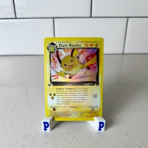 Może przedstawiać: Żółta karta Pokémon Dark Raichu z kreskówkową ilustracją postaci. Karta jest prezentowana na białym stojaku z niebieskimi literami "P". Tekst karty zawiera "Surprise Thunder" i "70 HP".