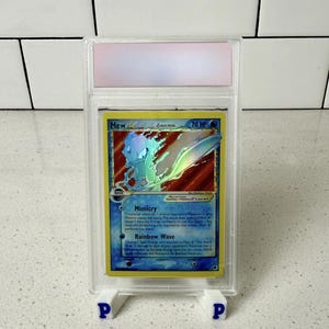 May include: A graded Pokémon card featuring Mew, a blue and purple creature, with the text "Mew" and "70 HP". The card is encased in a clear plastic holder, supported by a white stand with blue "P" letters.