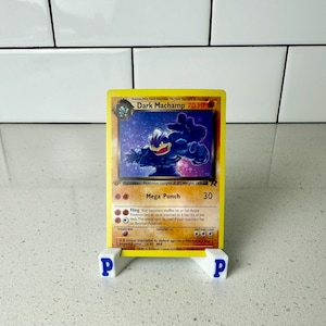 May include: A Dark Machamp Pokémon card, featuring a muscular, blue character with the text "Dark Machamp 70 HP." The card is displayed on a white stand with blue "P" letters. The card has a yellow border and a purple background.