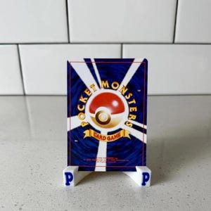 Op de afbeelding: Een Pok&eacute;mon-verzamelkaart op een witte standaard met blauwe letters "P". De kaart toont een rood-wit Pok&eacute; Ball-ontwerp met "Pocket Monsters Card Game" in goud op een blauwe achtergrond. De standaard staat op een wit oppervlak.