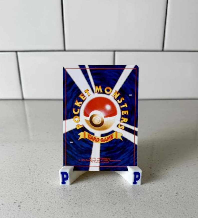 Op de afbeelding: Een Pok&eacute;mon-verzamelkaart op een witte standaard met blauwe letters "P". De kaart toont een rood-wit Pok&eacute; Ball-ontwerp met "Pocket Monsters Card Game" in goud op een blauwe achtergrond. De standaard staat op een wit oppervlak.