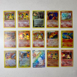 May include: A collection of fifteen Charizard Pokémon trading cards, featuring various designs and illustrations. The cards have yellow borders and include text and numerical values. Some cards have holographic elements.