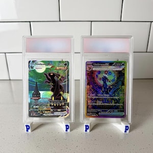 May include: Two Umbreon Pokémon trading cards in protective cases, displayed on white stands with blue "P" logos. The cards feature colorful artwork and text, including "Umbreon VMAX" and "Dark Signal". The background is a white tiled surface.