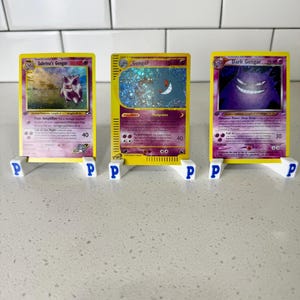 May include: Three collectible trading cards featuring Gengar characters, including Sabrina's Gengar, Gengar, and Dark Gengar. Each card is displayed upright in a small white plastic stand marked with the letter P, showcasing detailed artwork and text.