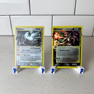 May include: Two Pokémon trading cards, Rayquaza and Charizard, displayed on white stands with blue "P" lettering. The cards have yellow borders and feature colorful illustrations of the Pokémon. The Rayquaza card shows a swirling blue and black design, while the Charizard card depicts a fiery orange and black creature.