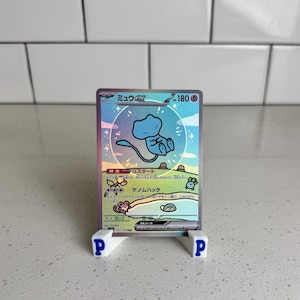 May include: A holographic Mew ex Pokémon trading card, featuring a blue Mew character. The card is displayed on a white stand with the letter "P" on each side. The card has Japanese text.