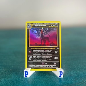May include: A Houndoom trading card with 70 HP, featuring a dark canine creature howling at a starry sky. The card displays the Dark Flame and Black Fang attacks, with text detailing its length of 4' 7" and weight of 77 lbs. It is held by a white stand.