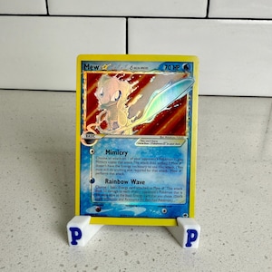May include: A holographic Mew Delta Species Pokémon trading card, displayed on a white stand with blue text. The card features a light purple and yellow creature with a blue and silver background.