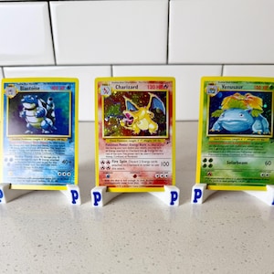 May include: Three Pokémon trading cards, Blastoise, Charizard, and Venusaur, are displayed on white stands with blue "P" logos. The cards have yellow borders and feature colorful illustrations of the Pokémon characters.