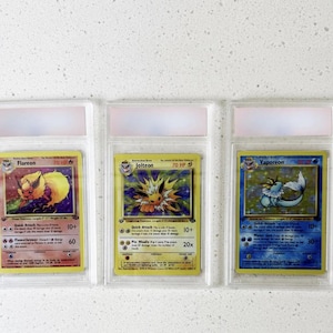 Może przedstawiać: Trzy karty kolekcjonerskie Pok&eacute;mon w ochronnych etui. Karty przedstawiają Flareona, Jolteona i Vaporeona, każda z unikalną grafiką i tekstem. Karty znajdują się w przezroczystych plastikowych etui z r&oacute;żowo-białą g&oacute;rą.