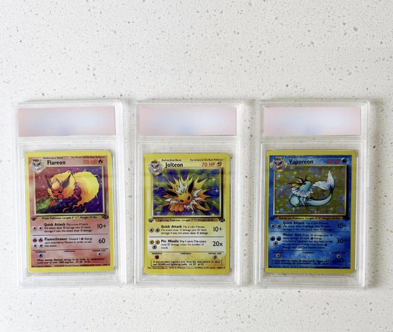 Może przedstawiać: Trzy karty kolekcjonerskie Pok&eacute;mon w ochronnych etui. Karty przedstawiają Flareona, Jolteona i Vaporeona, każda z unikalną grafiką i tekstem. Karty znajdują się w przezroczystych plastikowych etui z r&oacute;żowo-białą g&oacute;rą.