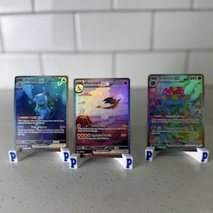 May include: Three Pokémon trading cards, featuring Blastoise, Charizard, and Venusaur, displayed on white stands with blue "P" logos. The cards have colourful illustrations and text, with a blue, orange, and green colour scheme. The stands are on a light-coloured surface.