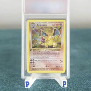 Peut inclure: Une carte à collectionner Charizard gradée, enfermée dans un étui de protection en plastique transparent, posée sur un support blanc avec des lettres P bleues. La carte montre une créature jaune, 120 PV, et les attaques Energy Burn et Fire Spin.