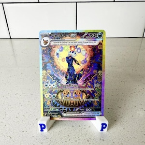 以下が含まれることがあります： 白いスタンドに青い「P」の文字が配置された、ホログラフィックUmbreon exポケモンカード。カードには、カラフルな背景と「Moon Mirage」のテキストが付いた、濃い青色のUmbreonキャラクターが描かれています。