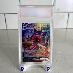 Puede incluir: Una carta coleccionable de Pokémon con Gengar VMAX, calificada y encapsulada en un soporte protector. La carta tiene una colorida ilustración de Gengar y el texto "Fear and Panic" y "G-Max Swallow Up". El soporte es blanco.