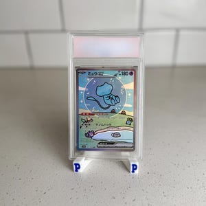May include: A graded Pokémon card featuring a blue Mew EX character, displayed in a clear plastic case. The card has Japanese text and is supported by two white stands with blue "P" letters.
