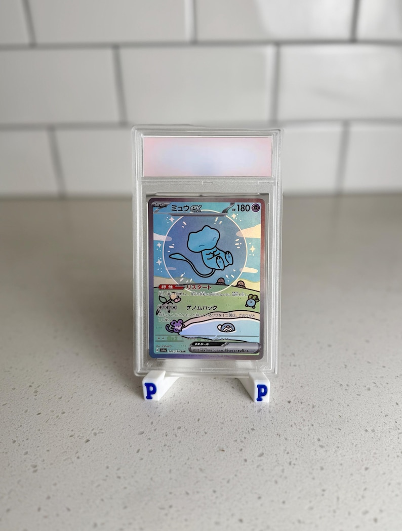 May include: A graded Pok&eacute;mon card featuring a blue Mew EX character, displayed in a clear plastic case. The card has Japanese text and is supported by two white stands with blue "P" letters.